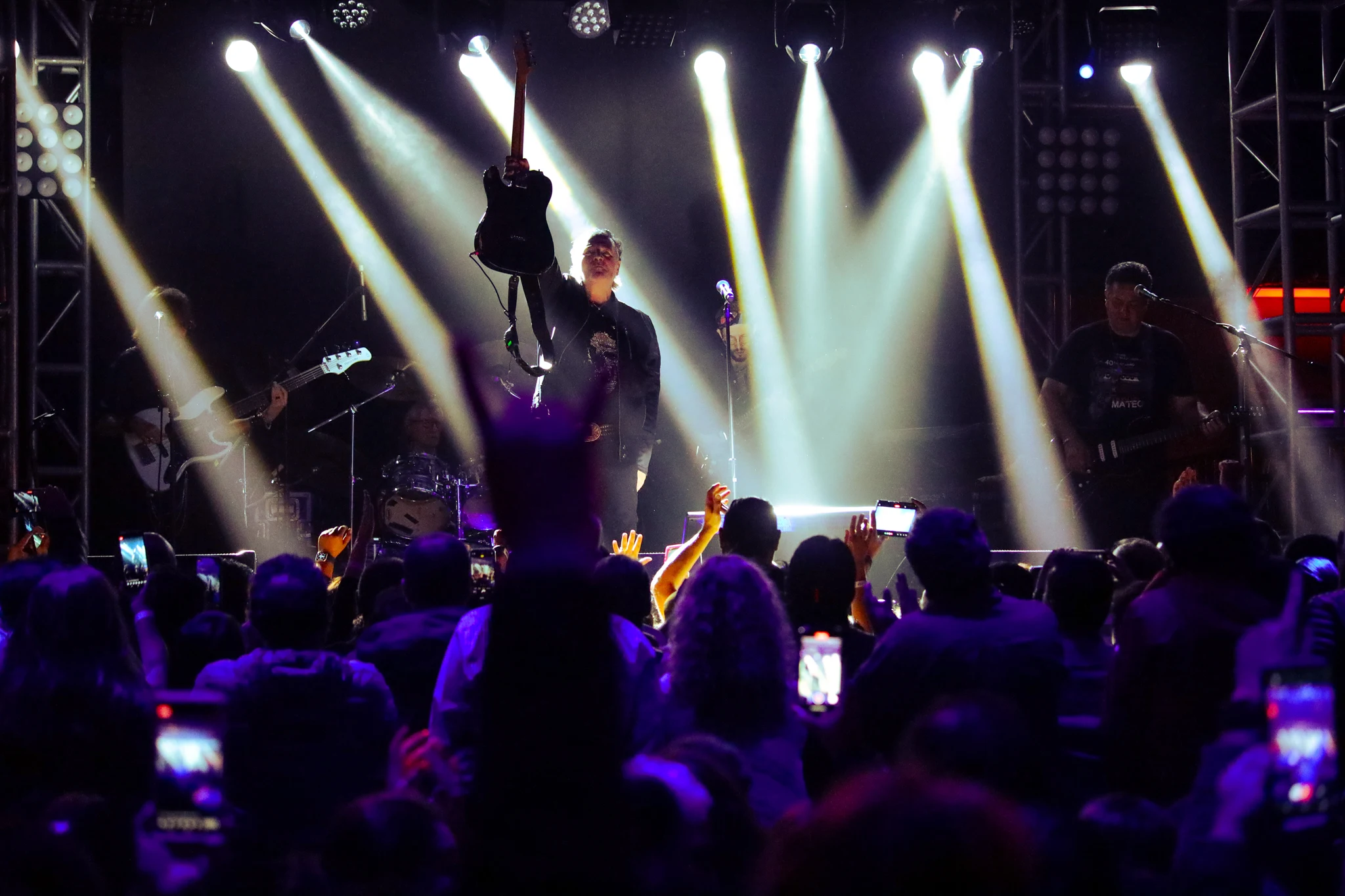 Guitarist performing under crossing spotlight beams as the crowd raises phones at a LIVEST show.
